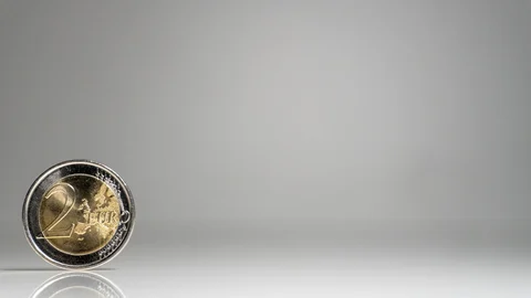 Moneda de 2 euros colocada de pie sobre una superficie reflectante y un fondo gris.