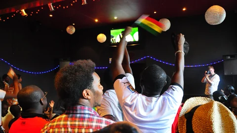 Aficionados al fútbol ven un partido televisado en un local y uno de ellos levanta una bandera de Ghana.
