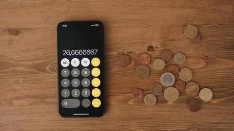 Un teléfono móvil con la calculadora abierta junto a varias monedas de euro sobre una mesa de madera.