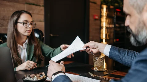 Dos personas intercambian un documento durante una reunión profesional alrededor de una mesa.