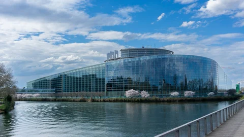 Edificio acristalado del Parlamento Europeo en Estrasburgo, junto al río, bajo un cielo parcialmente nublado.