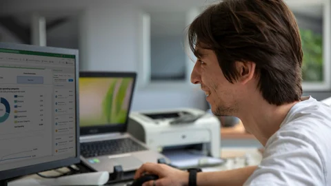 Joven trabajando frente a un ordenador con gráficos y datos en la pantalla, en una oficina.
