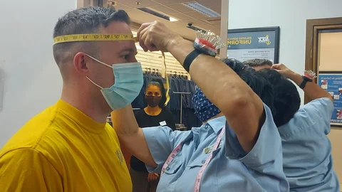 Un trabajador con mascarilla y camiseta amarilla mientras un empleado uniformado le mide la cabeza con una cinta métrica.