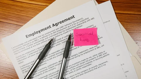 Contrato de trabajo sobre una mesa de madera, con dos bolígrafos y una nota rosa que dice «seasonal hire».